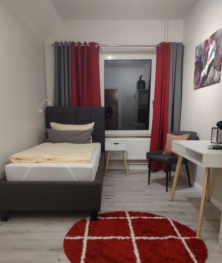 Das Bild zeigt ein Einzelzimmer mit einem Single-Bett auf der linken Seite, einem großen Fenster mit Vorhängen hinter dem Kopfteil und einem kleinen Schreibtisch mit Stuhl auf der rechten Seite. Auf dem Vinyl-Boden liegt dazwischen ein runder Teppich,