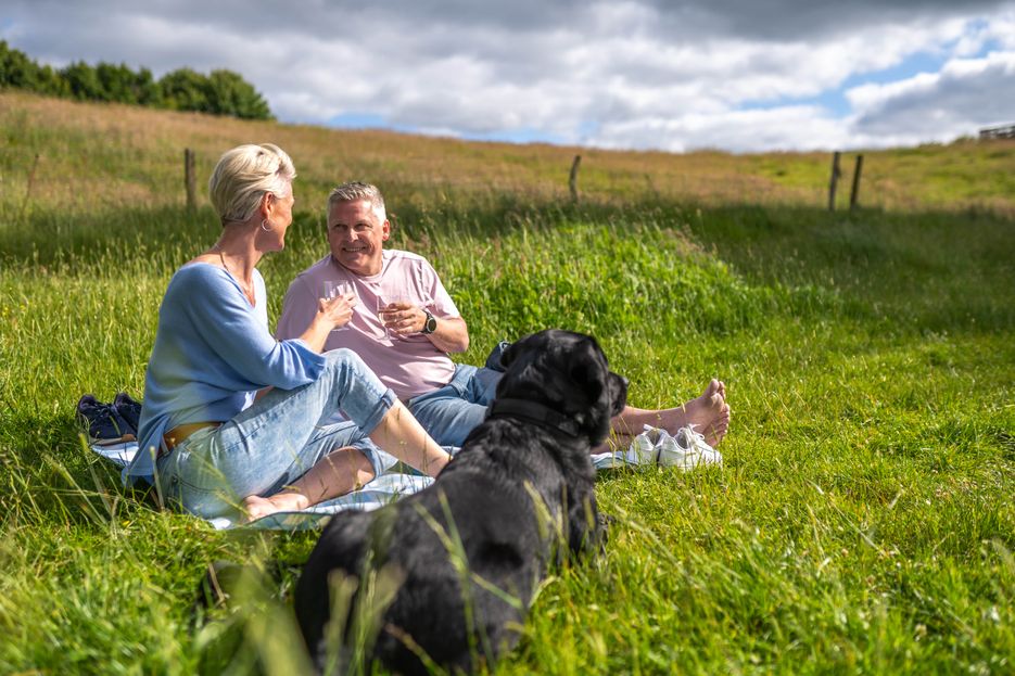 Zwei Menschen mit Hund sitzen im Gras und picknicken 