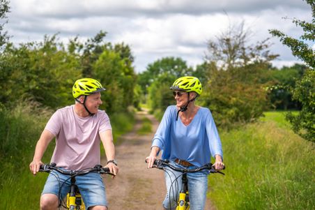 Ein Mann und eine Frau fahren lächelnd auf einem Feldweg Fahrrad. Beide tragen gelbe Helme und sommerliche Kleidung. Die Umgebung ist grün und ländlich.