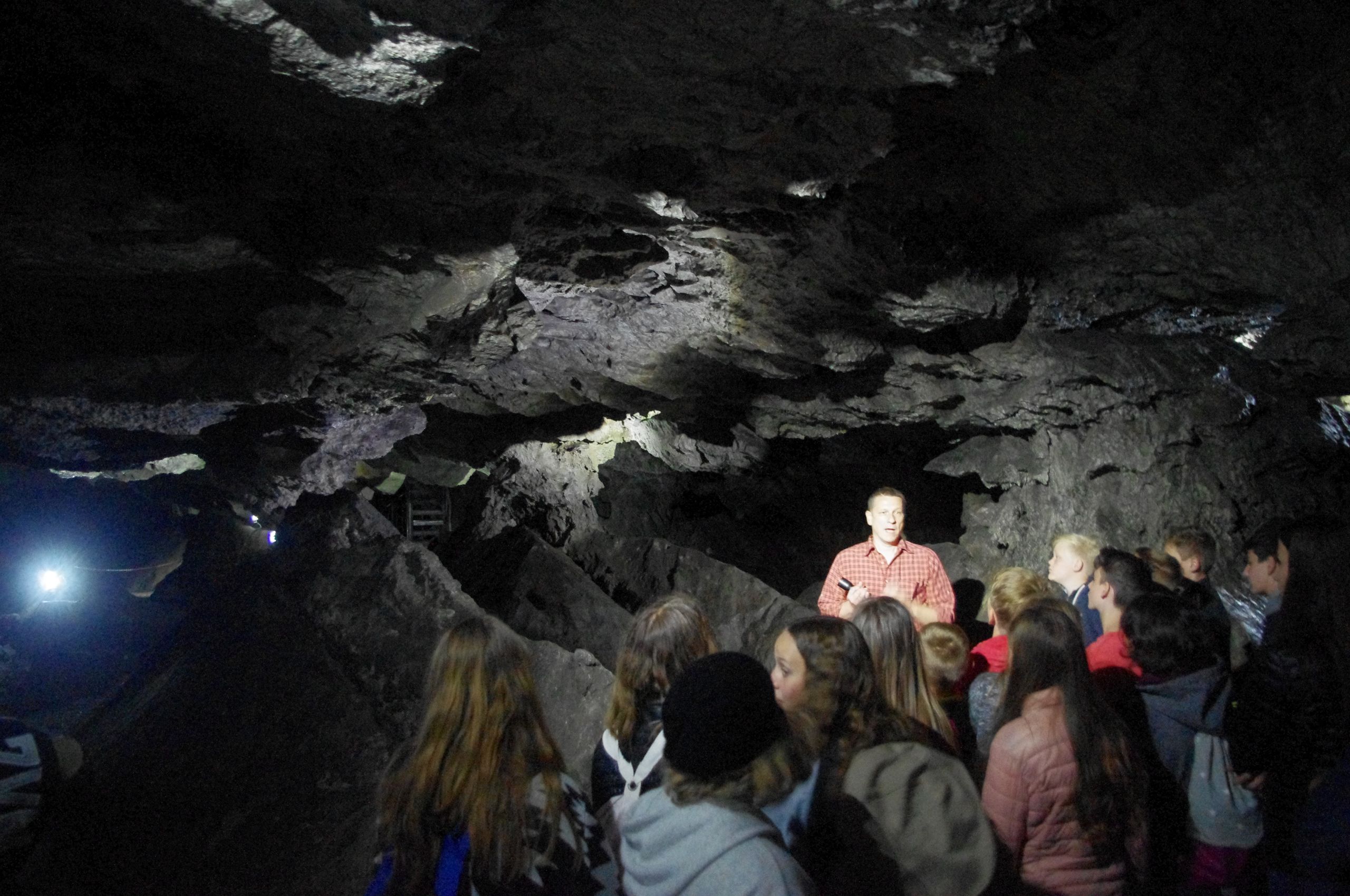 Eine Gruppe von Menschen hört einem Führer in einer dunklen Höhle zu. Der Führer trägt ein rotes Hemd und hält eine Taschenlampe.