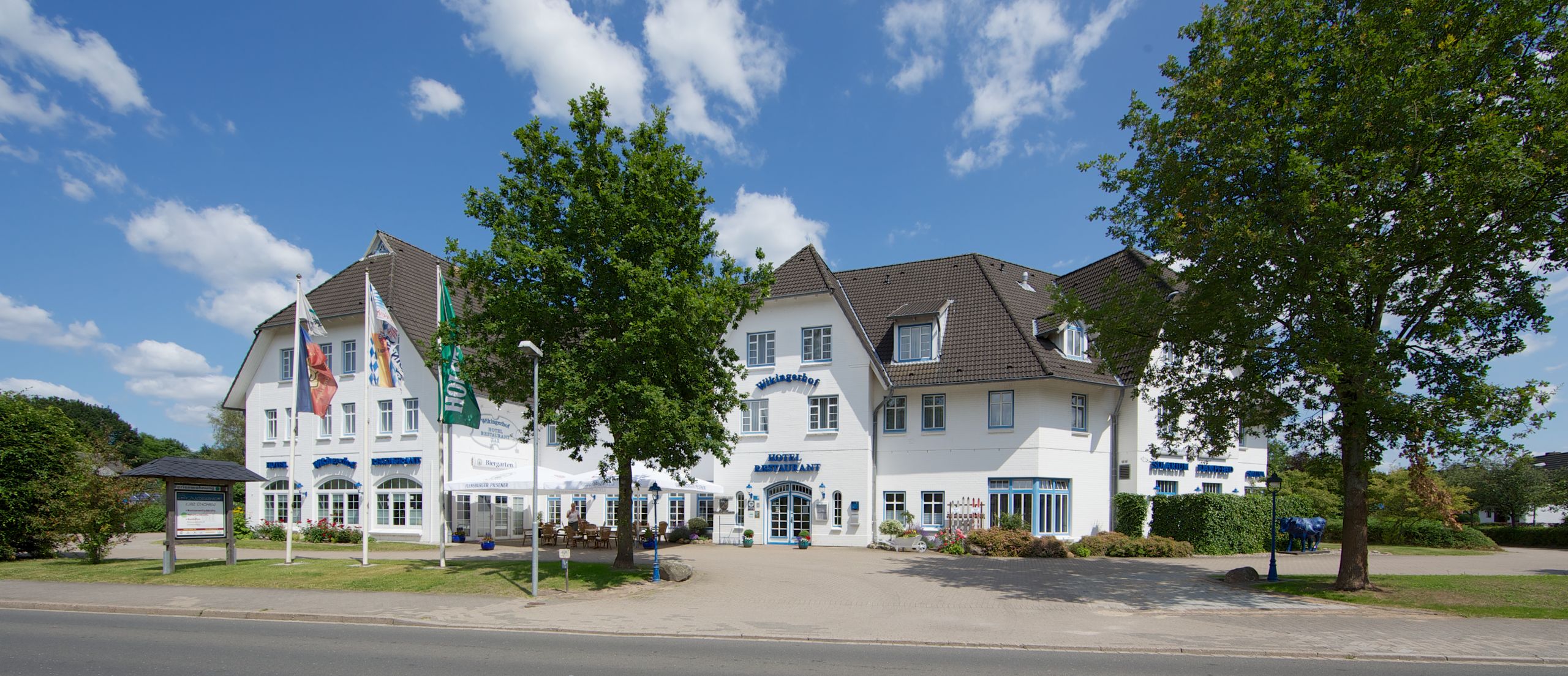 Das Hotel Wikingerhof zeigt sich mit weißer Fassade und mehreren Fahnen im Vordergrund. Ein Baum steht vor dem Gebäude, der Himmel ist blau mit wenigen Wolken.