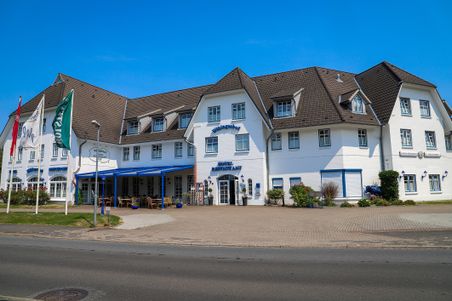 Das Hotel Wikingerhof zeigt sich in einem traditionellen Gebäude mit weißen Wänden und einem braunen Dach. Vor dem Hotel wehen Fahnen im Wind.