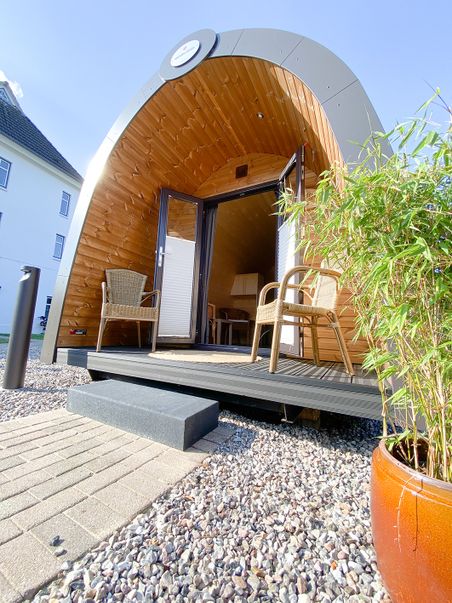 Moderne Lodge mit Holzverkleidung und Glasfront, umgeben von Kies und Pflanzen. Zwei Stühle stehen auf der Veranda, die Sonne scheint.