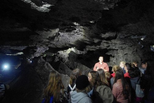 Eine Gruppe von Menschen hört einem Führer in einer dunklen Höhle zu. Der Führer trägt ein rotes Hemd und hält eine Taschenlampe.
