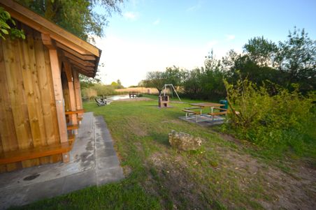 Holzhütte neben einem Grillplatz mit Picknicktischen und Schaukel. Im Hintergrund ein Steg am Wasser, umgeben von Bäumen und Wiesen.
