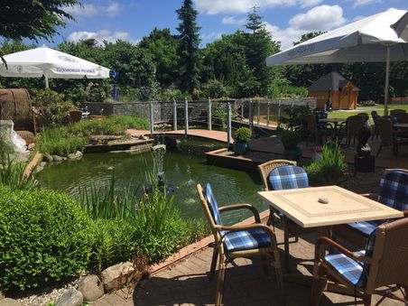 Ein idyllischer Biergarten mit Teich, Brücke und gemütlichen Sitzplätzen unter Sonnenschirmen. Perfekt für eine entspannte Auszeit im Freien.